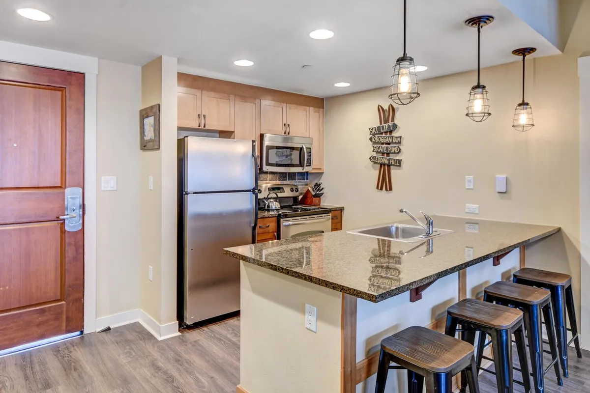 A welcoming kitchen scene featuring a granite countertop, stainless steel appliances, wooden bar stools, and tasteful wall art. The bar stools at the counter suggest casual dining, while the appliances and counter space highlight the kitchen’s functionality for both hosting and daily use.