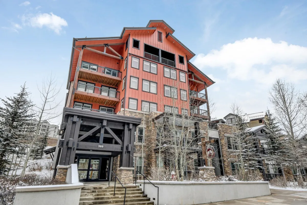 An inviting lodge-style entrance with a striking orange roof and stone façade, surrounded by winter scenery. Guests are seen relaxing inside, watching snowfall from balconies, and socializing by a cozy fire.