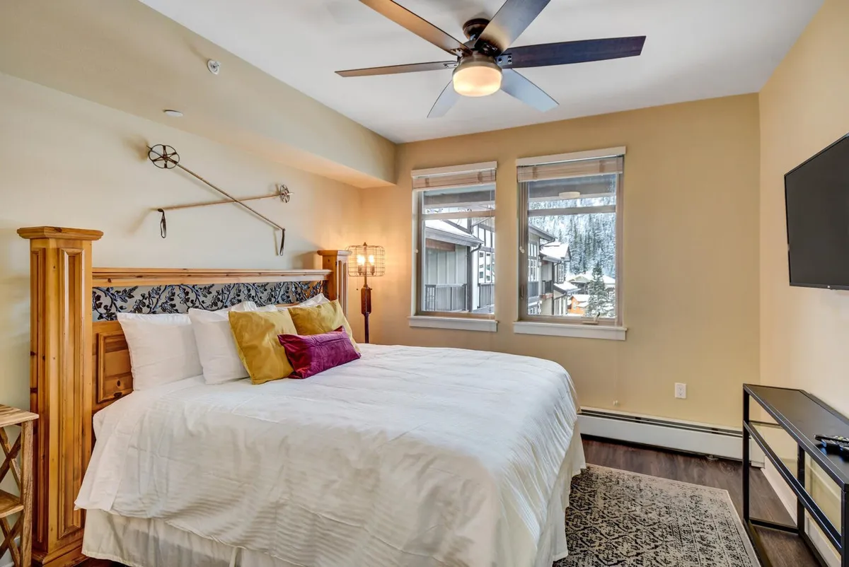 A tranquil bedroom featuring a large bed with colorful pillows, a wooden headboard, and windows that fill the space with natural light. The room exudes a peaceful atmosphere, ideal for relaxation and restful nights.