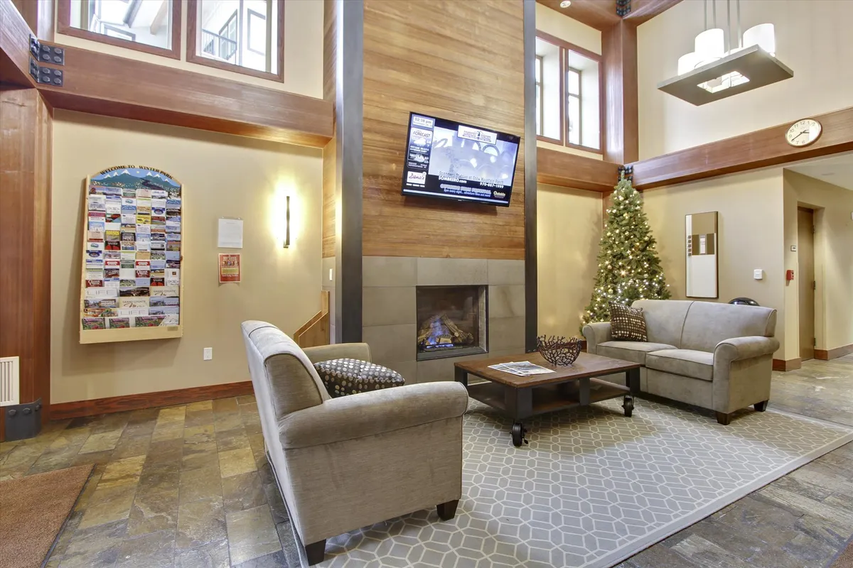 A stylish indoor lobby lounge featuring a cozy seating area by a modern fireplace, with a decorative tree and an information board nearby. The warm lighting and comfortable furnishings create a welcoming space for guests to relax, socialize, or gather information about resort activities.