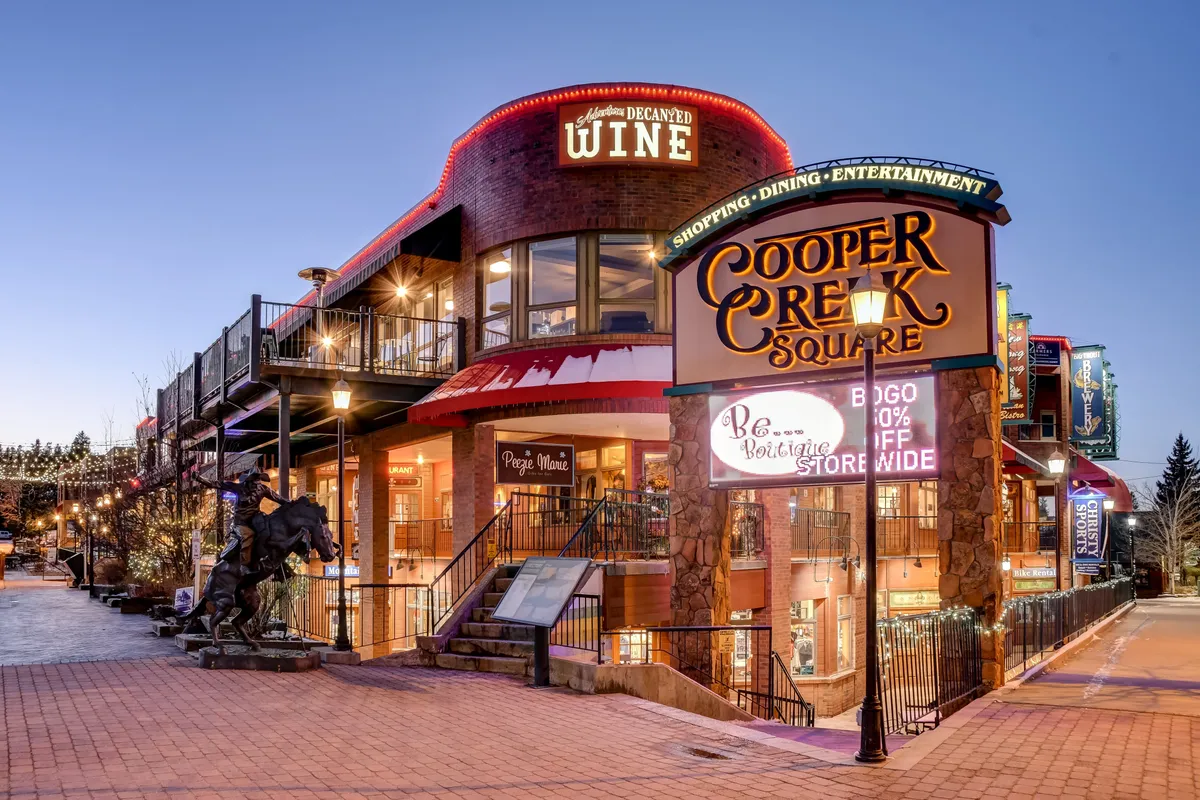 A lively outdoor shopping and dining area, featuring the Cooper Creek Square sign, wine bar signage, outdoor seating, and decorative lighting. The scene buzzes with the energy of local entertainment, culinary tastings, and social gatherings.