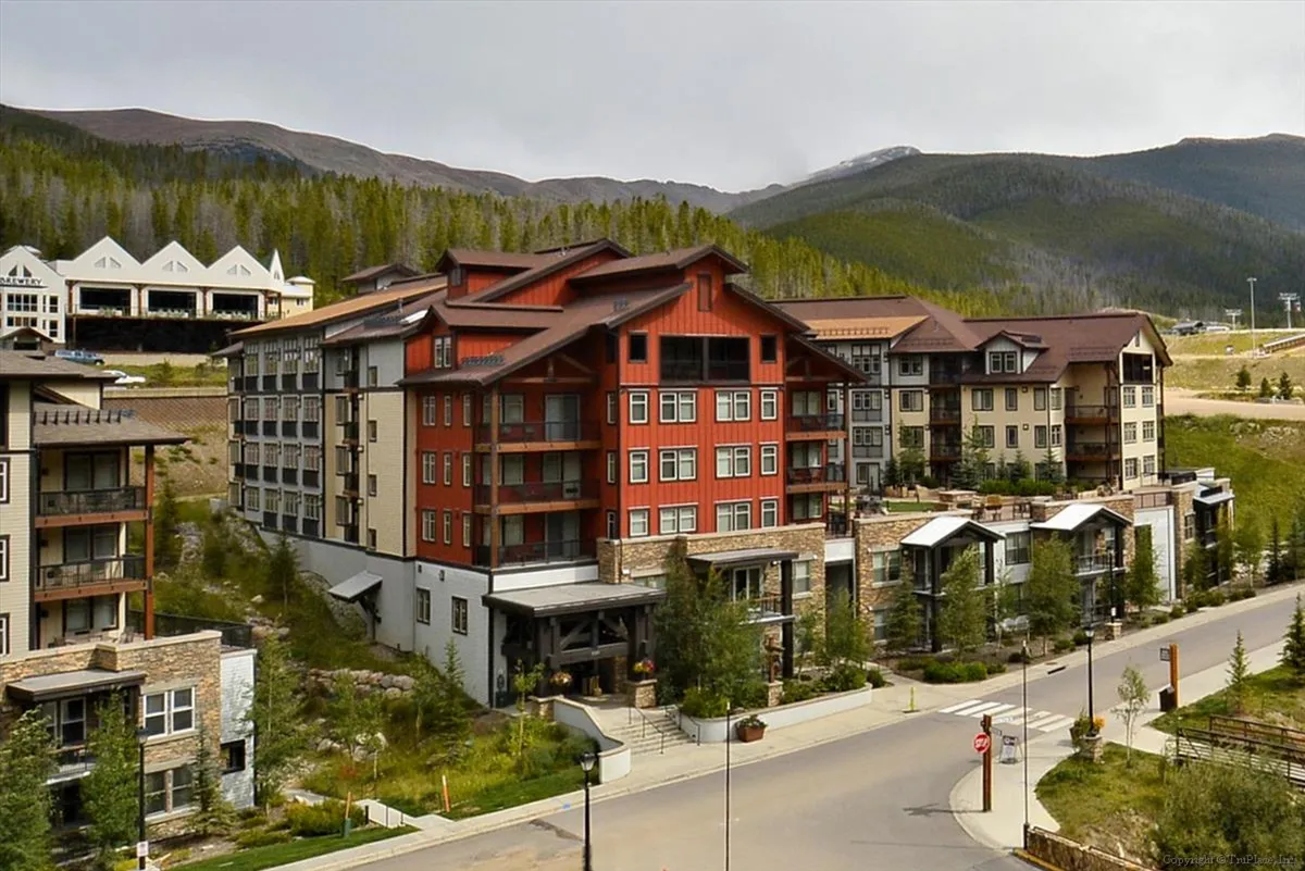 A lively outdoor scene showcasing modern resort architecture set against a mountain backdrop, surrounded by natural landscaping. Groups of people gather for community events and explore nearby trails, evoking an active and social atmosphere.