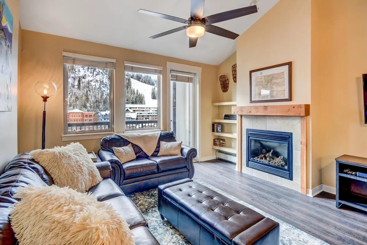 A cozy living room featuring a leather sofa arranged around a stone fireplace, with a large coffee table perfect for board games or shared snacks. Generous windows frame a scenic mountain view, while a modern HD Smart TV sits above the mantle, creating a blend of rustic charm and contemporary comfort.