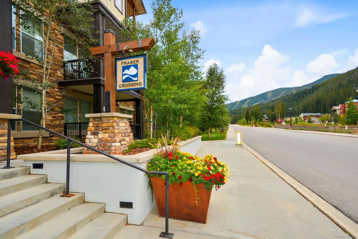 A welcoming exterior shot of the Fraser Crossing sign framed by colorful flower planters and stone steps. The scene suggests easy access to trails and the surrounding village, evoking the spirit of exploration and outdoor activity right outside the condo door.