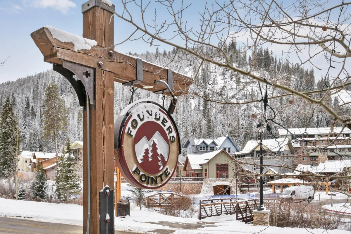 This exterior photo showcases the welcoming Founders Pointe sign surrounded by snow-covered trees, colorful buildings, and a dramatic mountain backdrop. The scene conveys the resort village atmosphere with its blend of natural beauty and vibrant community energy.