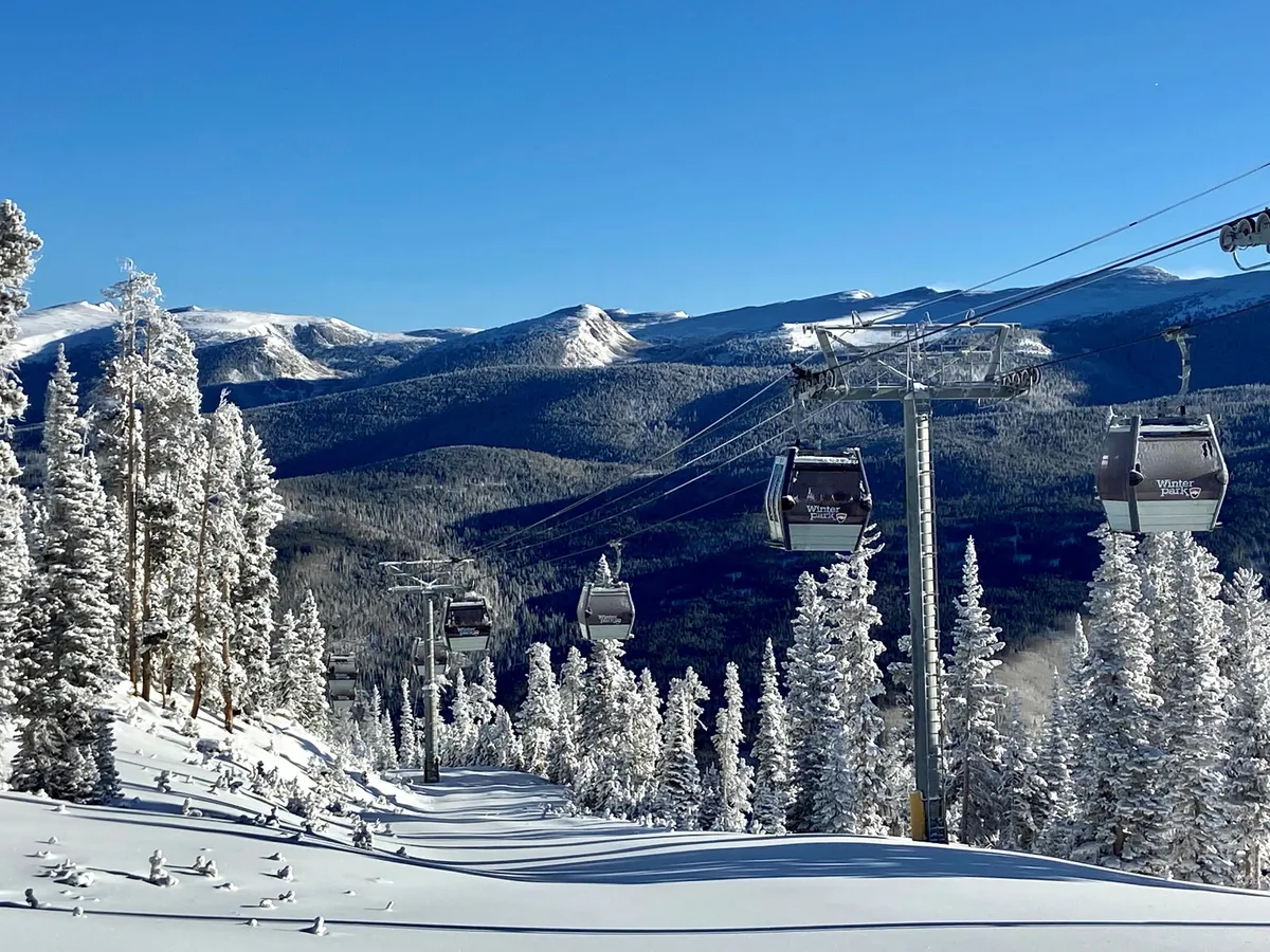 This image features ski lift gondolas gliding above snow-covered trees with a stunning mountain range in the background. The scene captures the wintry beauty and outdoor adventure possibilities of the Rocky Mountain region.