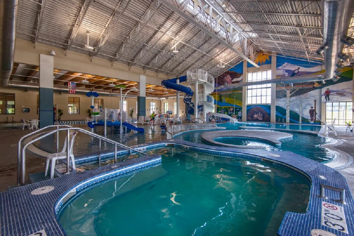 An indoor recreation center pool area featuring children playing, a colorful water slide, and vibrant murals. The joyful scene is filled with families enjoying swimming, water games, and celebrating special occasions by the pool.