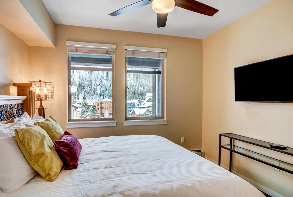 An inviting bedroom with a cozy bed adorned with colorful pillows, large windows offering mountain views, and a wall-mounted TV. The space blends comfort with modern amenities.