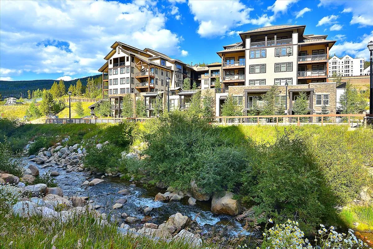 A tranquil scene showcasing modern building architecture beside a flowing stream, framed by lush greenery. The natural setting evokes relaxation and the connection between accommodation and nature.