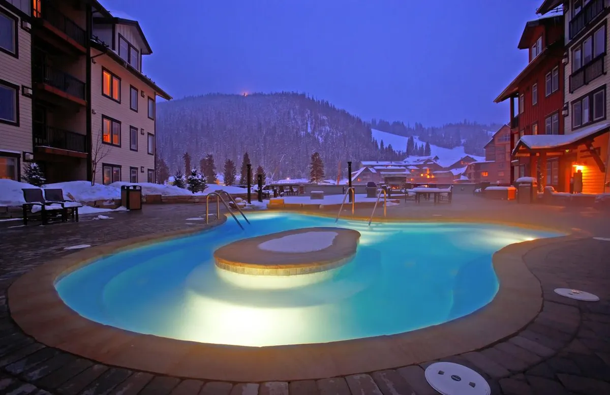 An outdoor hot tub illuminated against a snowy winter landscape, surrounded by warmly lit resort buildings. Several people are seen socializing in the steaming water while snow blankets the ground, creating a striking contrast between the cozy warmth of the tub and the chilly mountain environment.