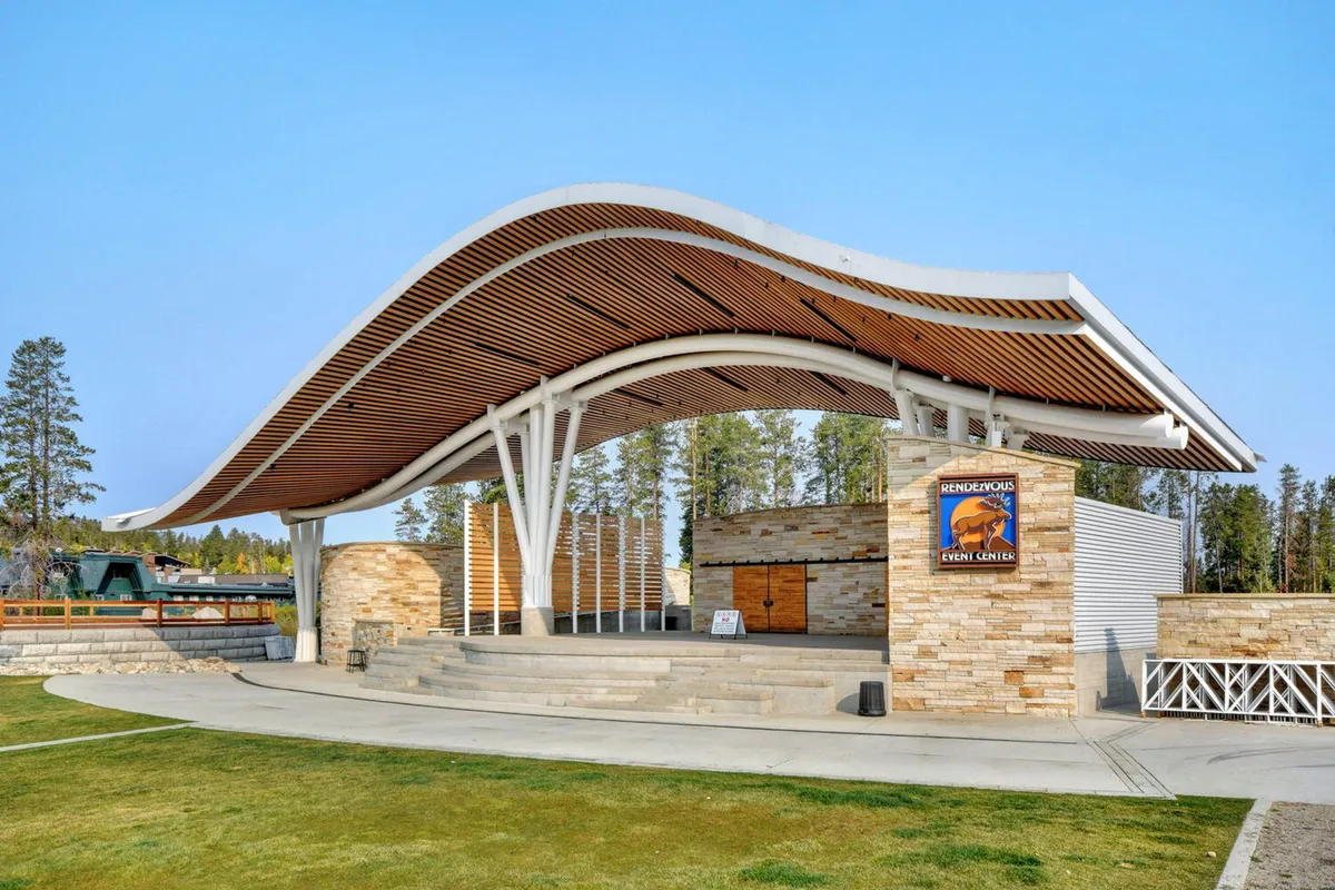 This exterior image features a distinctive venue with a curved roof structure, stone facade, and wooden elements, designed for concerts, community gatherings, and outdoor events. The scene suggests a space where memorable performances and celebrations take place, complementing the lively atmosphere of the area.