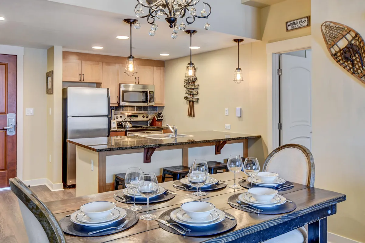 A beautifully set dining table is positioned in an open-concept kitchen, with modern appliances, an elegant chandelier, and decorative wall art visible. The table is ready for a shared meal, emphasizing opportunities for family gatherings or entertaining guests. The lighting and decor convey warmth and a sense of celebration.