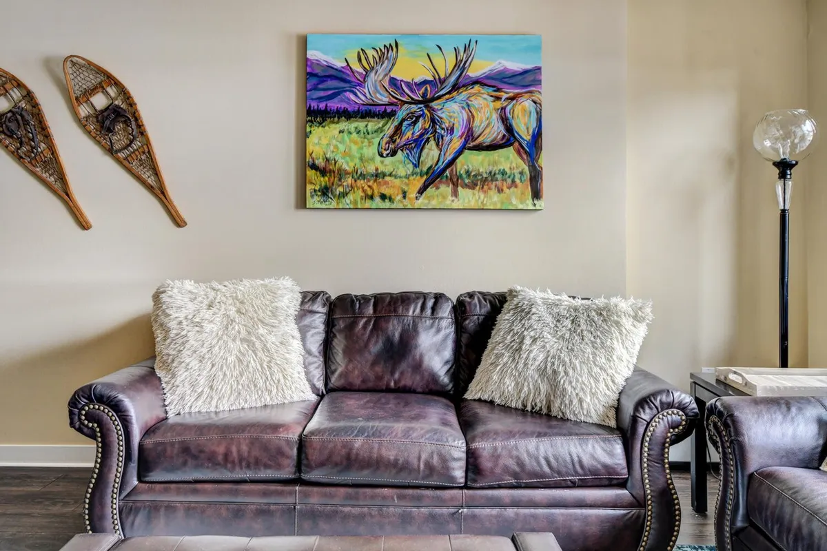 A stylish living area featuring a modern sofa (ideal as a sleeper sofa), a colorful moose painting, decorative snowshoes, and a modern lamp. The setting is cozy and artistic, suited for both relaxation and socializing.