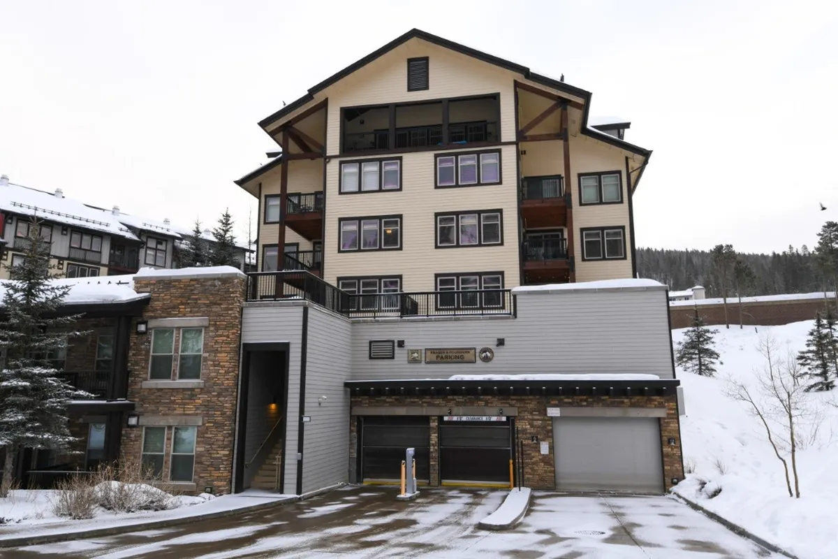 A multi-story building with a stone facade, balconies, and a snow-covered ground, situated in a vibrant winter setting. Guests can be seen gathering and enjoying the wintry landscape, with the promise of skiing and local shops nearby.
