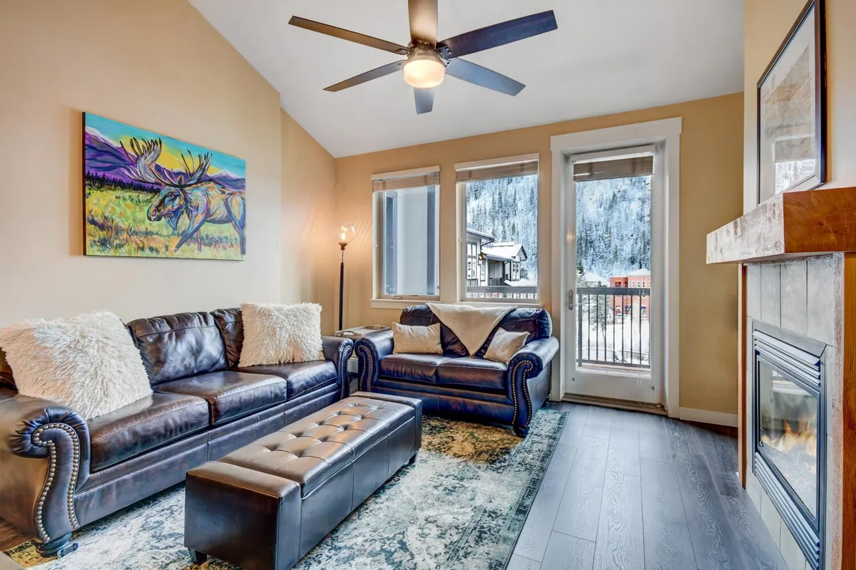 The image depicts a welcoming living room featuring plush leather sofas centered around a glowing fireplace, with striking local artwork on the walls and expansive windows that offer breathtaking views of snow-dusted mountain peaks. Soft natural light fills the space, creating a cozy atmosphere perfect for relaxing after a day on the slopes.